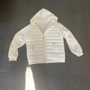 Vit cardigan med huva - Snygg vit cardigan med quiltade partier och dragkedja framtill. Jackan har huva, två fickor med dragkedja och ribbade muddar. Perfekt för dig som gillar en clean och stilren look. Materialet känns mjukt mot huden. Har aldrig varit utomhus med den. (Gratis frakt till 19:e september!!)