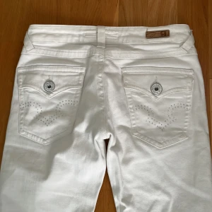 Vita jeansbyxor från Gina Tricot - Snygga vita utsvängda jeansbyxor från Gina Tricot med silverfärgade knappar och nitar. Bakfickorna har dekorativa sömmar och små strassdetaljer. Klassisk femficksmodell i stretchig bomullsmix, perfekt för en fräsch look.❤️De är lite smutsiga på slutet av ena byxbenet och har en ljusare fläck där fram som jag kan skicka privat då jag inte fick plats med fler bilder i annonsen😅 Midjemått rakt över: 36 och innerbenslängden: 79cm❤️