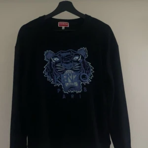 Svart Kenzo tröja med tigerbrodyr - Svart tröja från Kenzo med ikonisk blå och lila tigerbrodyr framtill och texten 'Kenzo Paris'. Tröjan har rund halsringning och långa ärmar. Perfekt för dig som vill sticka ut med en snygg streetstyle-look.