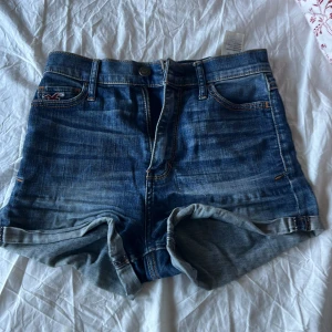 Blå jeansshorts från Hollister - Snygga blå jeansshorts från Hollister med klassisk femficksdesign, uppvikta benslut och hög midja. Shortsen har slitna detaljer och tydliga sömmar med orange tråd. Perfekta för varma dagar och enkel att matcha med allt.