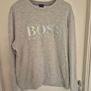 Grå sweatshirt från Hugo Boss - Snygg ljusgrå sweatshirt från Hugo Boss med stort logotryck framtill. Tröjan har rund hals, långa ärmar och ribbade muddar. Slim fit-modell i mjuk bomullsblandning, perfekt för en clean och stilren look.