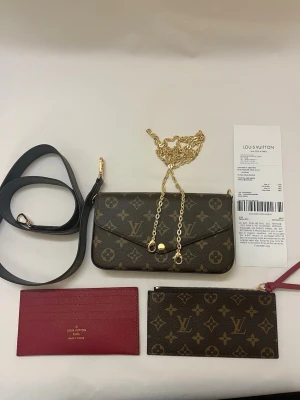 Louis Vuitton Félicie Pochette Flap Pouch - Säljer en ikonisk Louis Vuitton axelväska i klassiskt monogrammönster. Väskan har guldfärgad kedja, avtagbar svart axelrem och matchande monogram-plånbok samt röd kortficka. Materialet är exklusivt skinn med LV-logga och detaljer i guld.