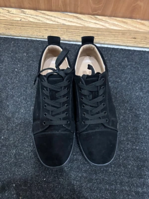 Svarta mockasneakers från Louboutin - Säljer ett par svarta sneakers från Louboutin i mjuk mocka med klassisk röd sula och svarta skosnören. Skorna har rund tå, platt sula och diskreta detaljer som metallöljetter och dragflik bak. Insidan är fodrad i beige skinn för extra komfort.