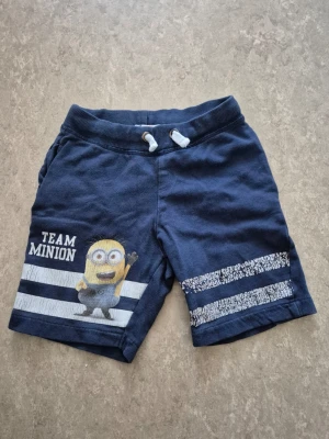 Shorts - Mörkblå shorts från H&M med Team Minion-tryck och en Minion-figur på ena benet. Shortsen har vita snören i midjan och två vita ränder på båda benen.