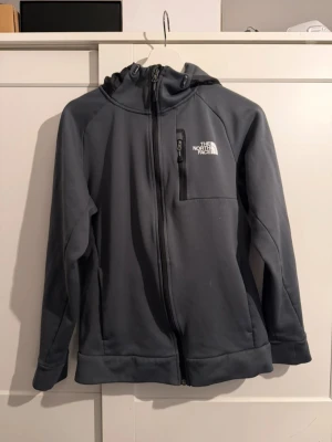 Mörkgrå vindjacka från The North Face - Säljer en mörkgrå vindjacka från The North Face med huva och dragkedja framtill. Jackan har en bröstficka med dragkedja och logga på både fram- och baksidan. Tillverkad i mjukt syntetmaterial och passar perfekt för blåsiga dagar.