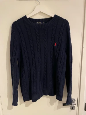Mörkblå kabelstickad tröja Polo Ralph Lauren - Kabelstickad mörkblå tröja från Polo Ralph Lauren med klassisk röd logga broderad på bröstet. Tröjan har rund halsringning, ribbade muddar och lång ärm. Perfekt plagg nu till vintern. Storlek L men passar M utmärkt