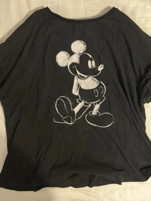Svart Mickey Mouse t-shirt Disney - Svart pyjamas t-shirt från Disney med en stor vit skiss av Mickey Mouse på framsidan. T-shirten har korta ärmar och en lös passform, perfekt för dig som gillar en chill och avslappnad stil. Tillverkad i mjuk bomull för extra komfort.