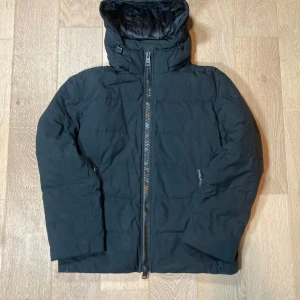Herno jacka - Säljer en svart pufferjacka med huva och hel dragkedja framtill. Jackan har en quiltad look och två sidofickor med dragkedja. Perfekt för kalla dagar när du vill hålla dig varm och stilsäker.