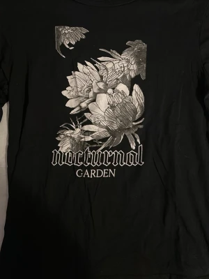 Svart t-shirt Nocturnal Garden X - Svart t-shirt från 157 The Black Label Collection i storlek XS. Framsidan har ett stort tryck med blommor och texten 'nocturnal garden' i vit/grafit. Klassisk rund hals och korta ärmar. Perfekt för dig som gillar edgy och stilrena prints.