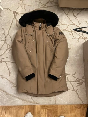 Moose Knuckles brun dunjacka med päls - Säljer en brun dunjacka från Moose Knuckles med svart pälskantad huva storlek m men passar som L/XL