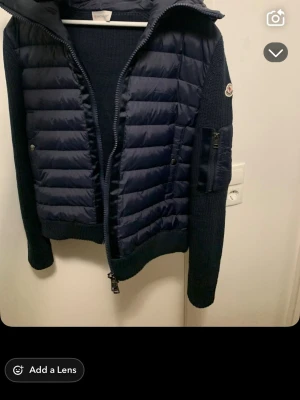 Mörkblå pufferjacka från Moncler - Säljer en mörkblå pufferjacka från Moncler med stickade ärmar och quiltad kropp. Jackan har dragkedja framtill, ficka med dragkedja på ärmen och en klassisk Moncler-logga på armen. Perfekt för dig som vill ha en snygg och sportig look.