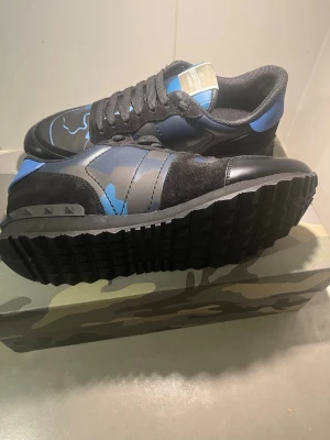 Valentino Garavani Rockrunner sneakers - Säljer ett par Valentino Garavani Rockrunner sneakers i svart, blått och grått med camouflagemönster. Skorna har snörning, detaljer i mocka och skinn samt en robust svart sula. Ikonisk Valentino-logga på plösen och coola nitar på hälen.