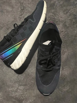 Louis Vuitton svarta sneakers med regnbågsdetalj - Säljer ett par svarta Louis Vuitton sneakers med stickad ovandel och snörning. Skorna har en vit, chunky sula och cool regnbågsfärgad detalj på sidan samt LV-logga i lila bak. Perfekta för dig som vill ha något unikt och trendigt i garderoben.