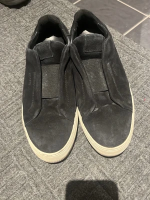 Svarta slip-on sneakers från Axel Arigato - Säljer ett par svarta slip-on sneakers från Axel Arigato i mocka med elastiska band över vristen. Skorna har en vit platt sula och minimalistisk design med diskret logga på sidan. Perfekta för dig som gillar stilrena och bekväma skor.