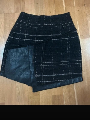 Svart kjol  från Zara Basic S - Unik svart kjol från Zara Basic i storlek S. De har rutigt mönster, asymmetrisk omlottfront och detaljer i skinnimitation. Dragkedja bak och snygg struktur i tyget. Perfekt för dig som vill ha något annorlunda och trendigt.