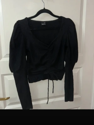 Svart långärmad topp med snörning - Säljer en svart långärmad topp från Ginatricot med v-ringning och puffiga axlar. Toppen har snygg snörning framtill som ger en croppad look och är gjord i ett mjukt, stickat material. Perfekt att styla med höga jeans eller kjol.