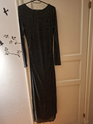 Svart glittrig långklänning från new yorker - Supersnygg svart långklänning  med glittriga prickar över hela tyget. Klänningen har lång ärm och en djup, rundad rygg. Perfekt för dig som vill sticka ut med en elegant och trendig look. Materialet känns mjukt och följsamt mot kroppen.