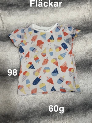 Färgglad glassmönstrad t-shirt - Söt t-shirt med glassar i olika färger och former över hela plagget. Basen är vit och trycket går i blått, rött, gult och orange. T-shirten har korta ärmar och rund halsringning med vit kant. Perfekt för sommarens alla äventyr!🌸 Fläckar. Storlek 98