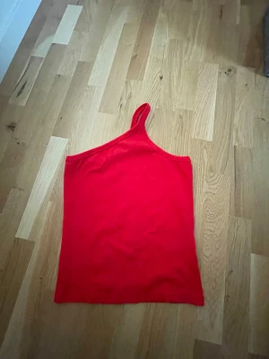 Röd oneshoulder topp - En ribbad och snygg röd oneshoulder topp. Toppen har en axelbandslös design på ena sidan och ett smalt axelband på andra sidan. Oanvänd!