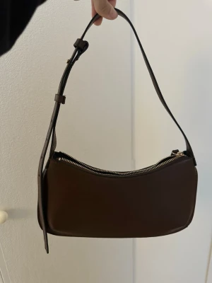 Brun axelväska i skinn - Säljer en stilren brun axelväska i skinn med justerbart axelband och snygg guldfärgad dragkedja. Väskan har en minimalistisk design och passar perfekt till alla dina outfits. Perfekt storlek för det viktigaste och enkel att bära över axeln.