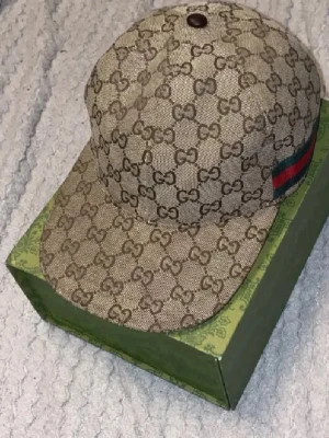 Beige Gucci keps med GG-mönster - Säljer en beige keps från Gucci med klassiskt GG-mönster över hela kepsen och den ikoniska gröna och röda randen på sidan. Kepsen är tillverkad i ett slitstarkt material och har mörkbrunt innerfoder. Perfekt accessoar för dig som gillar lyxiga detaljer.