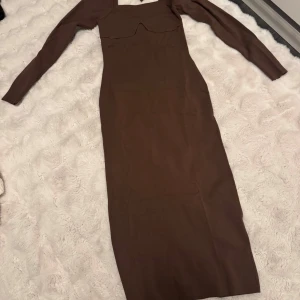 Brun långärmad klänning med knyt - Säljer en brun långklänning med långa ärmar och snygg knytning i nacken. Klänningen har en unik detalj vid bysten och är gjord i ett stretchigt, ribbat material som sitter snyggt på kroppen. Perfekt för dig som gillar stilrena och trendiga plagg.