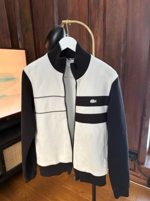 Svartvit ziptröja från Lacoste - Säljer en clean svartvit ziptröja från Lacoste med ribbad krage och muddar. Tröjan har svarta ärmar, vita paneler framtill och två svarta ränder med Lacoste-logga på bröstet. Regular fit och tillverkad i bomullsmix. Perfekt till lager på lager under höst och vår. Finns en flaw som syns på bild 2 inget som är märkbart synligt vid användning 