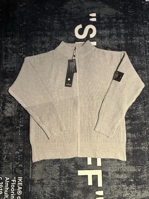 Grå stickad kofta från Stone Island - Säljer en grå stickad kofta från Stone Island med ribbad struktur och dragkedja framtill. Klassisk logga på vänster ärm och hög krage. Perfekt för lager-på-lager under kyliga dagar. Enkel och stilren design som passar till det mesta.