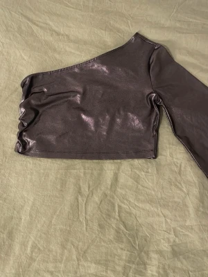 Glittrig one shoulder topp från NA-KD - Säljer en svart glittrig one shoulder topp från NA-KD. Toppen har lång ärm på ena sidan och är axelbandslös på andra. Materialet är glansigt och stretchigt och det är en magtröja.