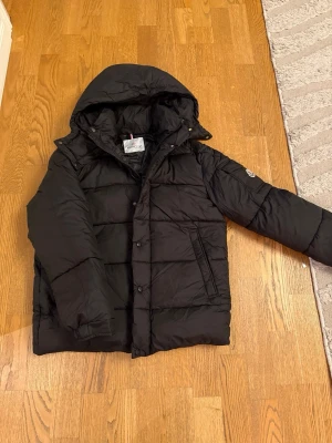 Moncler Maya 70 Hooded Short Down Jacket - Säljer en svart pufferjacka från Moncler med huva och klassisk logga på ärmen. Jackan har dragkedja och knappar framtill, samt fickor på sidorna och en extra ficka på ärmen. Insidan har ett coolt serietidningsmönster. Perfekt för kalla dagar.