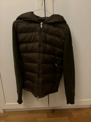 Mörkgrön Moncler pufferjacka med huva - Säljer en comprehensive mörkgrön pufferjacka från Moncler med stickade ärmar och vadderad kropp. Jackan har dragkedja framtill, huva med broderad logga och en Moncler-patch på ärmen. Perfekt för kyliga dagar och riktigt snygg till streetwear.