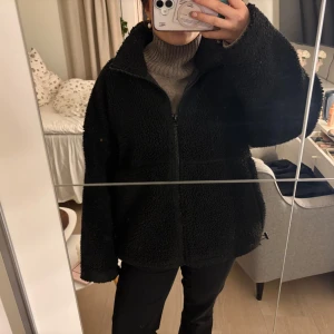 Svart teddyjacka med dragkedja - Mysig svart teddyjacka med hög krage och hel dragkedja framtill. Jackan har en loose passform och är tillverkad i fluffigt teddy-material som håller dig varm. Perfekt för kyliga dagar och enkel att styla till olika outfits.