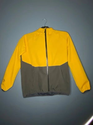 Gul och grå regnjacka från Neomondo - Snygg tvåfärgad regnjacka från Neomondo i storlek 164. Jackan är gul upptill och på ärmarna, med grå nederdel och rygg. Den har huva, hel dragkedja framtill och elastiska muddar vid ärmslut och midja. Perfekt för blåsiga dagar.