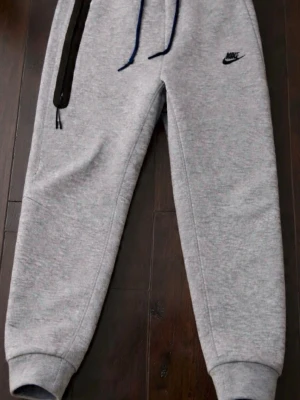 Grå mjukisbyxor från Nike - Snygga grå mjukisbyxor från Nike med svart logga på låret, svarta dragkedjor på ena fickan och blå snörning i midjan. Perfekta för chill eller träning, med mjukt material och muddar vid bensluten.