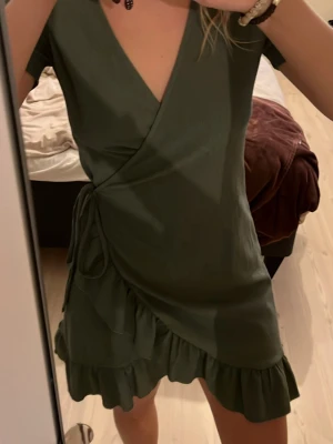 Grön omlottklänning från Vero Moda - Snygg grön omlottklänning från Vero Moda med volangkant nertill och knyt i sidan. Klänningen har korta ärmar och v-ringad hals. Perfekt för dig som gillar en feminin och enkel stil.