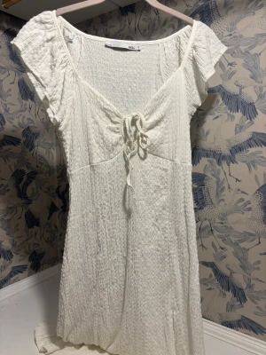 Vit långklänning med snörning 157 - Supersöt vit kortklänning från Lager157 med strukturmönstrat tyg och snörning framtill. Klänningen har korta ärmar och en lätt v-ringad halsringning. Perfekt för varma dagar och enkel att styla med accessoarer.