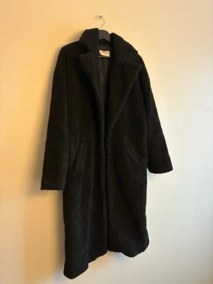 Svart lång teddyjacka  - Mysig svart teddyjacka från Na-kd i lång modell. Jackan har stora slag, sidofickor och är helfodrad för extra värme. Perfekt för kalla dagar och ger en cool oversized look.