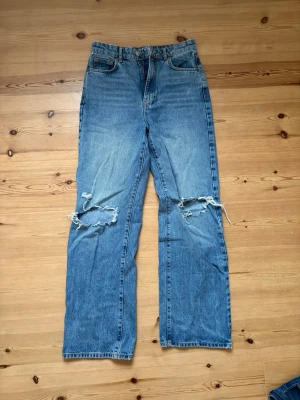 Blå jeans med hål - Jeans i ljusblå tvätt med raka ben och hög midja. Jeansen har tydliga slitningar och hål över båda knäna. Från Gina Tricot i storlek 38. 