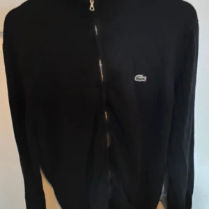 Svart kofta från Lacoste med dragkedja - Säljer en svart kofta från Lacoste i regular fit med hel dragkedja framtill och den klassiska krokodilloggan på bröstet. Tröjan har ribbade muddar och lång ärm, perfekt för dig som gillar stilren och enkel design.
