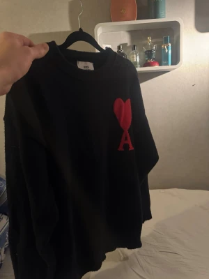 Ami tröja storlek M - Säljer en svart stickad tröja från Ami Alexandre Mattiussi med röd hjärt- och A-detalj på bröstet. Tröjan har rund halsringning, ribbade muddar och lång ärm. Perfekt för dig som vill ha en stilren men ändå unik look.