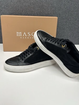 Svarta sneakers från Mason Garments - Snygga svarta sneakers från Mason Garments med ovandel i mocka och skinn, vita gummisulor och guldfärgade metalldetaljer vid snörningen. Klassisk låg modell med rund tå och Mason-logga på hälen. Perfekta för dig som gillar stilrena och exklusiva sneakers. Ny pris 3600kr. 100% äkta. Skriv för frågor