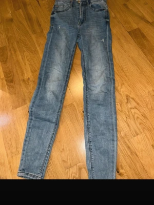 Blå skinny jeans från Stradivarius - Säljer ett par ljusblå skinny jeans från Stradivarius i storlek 34. Jeansen har hög midja, klassisk femficksdesign och dragkedja. De har slitna detaljer på låret och knät för en trendig look. Perfekta till sneakers eller boots.