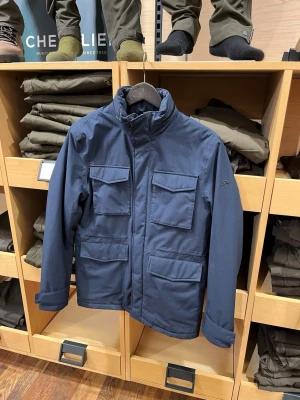 Blå fältjacka från J.Lindeberg - Snygg mörkblå fältfrån J.Lindeberg med fyra stora fickor framtill och hög krage. Jackan har en stilren look med diskret logga på ärmen och stängs med dragkedja. Perfekt för dig som vill ha en praktisk och clean jacka till vardags. Den är dunfodrad