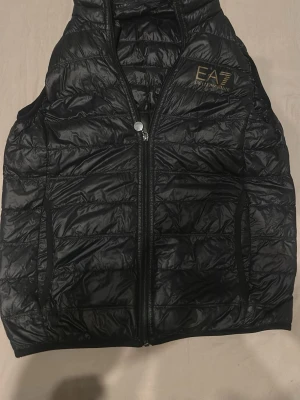 Svart dunväst från EA7 Emporio Armani - Snygg svart dunväst från EA7 Emporio Armani med guldfärgad logga på bröstet. Västen har quiltad design, dragkedja framtill och två sidofickor. Perfekt att slänga över en hoodie eller tröja för en sportig look.