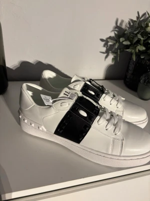 Valentino Rockstud Untitled sneakers - Säljer ett par vita Valentino Rockstud Untitled sneakers med svart läderdetalj och svarta nitar. Skorna har klassisk snörning, platt sula och coola pyramidformade nitar längs sidan och på sulan. Märkeslogga syns både på plösen och innersulan. Perfekta för dig som gillar statement-detaljer.