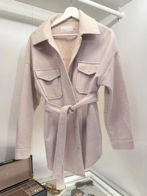 Beige overshirt/kalla - Nelly  - Snygg oversized beige overshirt/kappa med bälte i midjan från NLY Trend. Jackan har bröstfickor och svarta knappar som ger en snygg kontrast, klassisk krage och långa ärmar. Plagget är endast använt ett fåtal gånger och är i mycket fint skick. Eftersom att modellen är ”oversized” så sitter jackan som en storlek S/M. Hör av dig om du vill se hur den sitter på! 