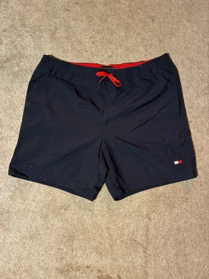 Marinblå badshorts Tommy Hilfiger - Säljer ett par marinblå badshorts från Tommy Hilfiger med röd dragsko och meshfoder. Shortsen har en liten logga fram och en stor flaggdetalj bak på fickan. Perfekta för stranden eller poolen!