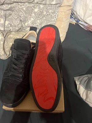Svarta sneakers med röd sula från Louboutin - Säljer ett par svarta sneakers från Christian Louboutin med ikonisk röd sula och snygg snörning. Ovandelen är i mocka och textil, och insidan är ljus. Perfekta för dig som vill sticka ut med exklusiva detaljer och klassisk design. ALLTING MEDKOMMER!