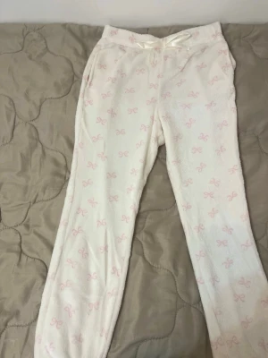 Vita mjukisbyxor med rosa rosetter - Supermysiga vita mjukisbyxor med ljusrosa rosett-mönster och en söt satinrosett i midjan. Byxorna har normal passform, resår i midjan och är gjorda i ett fluffigt, mjukt material som känns riktigt skönt mot huden.