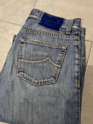Blå jeans från Jacob Cohen, storlek 32 - Säljer ett par klassiska blå jeans från Jacob Cohen i storlek 32. Jeansen har raka ben, fem fickor och snygga kontrastsömmar. Märkespatch i blå sammet baktill och slitning på vänster lår för en cool look. Perfekta till vardagsoutfiten.